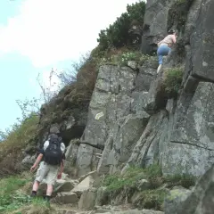 Turysta z plecakiem wspina się po skalistej ścieżce, obok wspina się inna osoba.