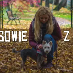 Kobieta z psem na tle jesiennego parku, pokrytego liśćmi.