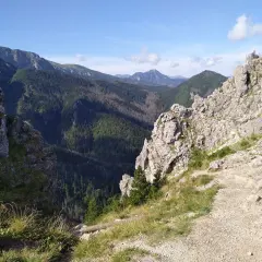 Widok na skaliste szczyty i zalesione doliny Beskidu Wyspowego. Na jednym ze szczytów siedzi człowiek, podziwiając panoramę.