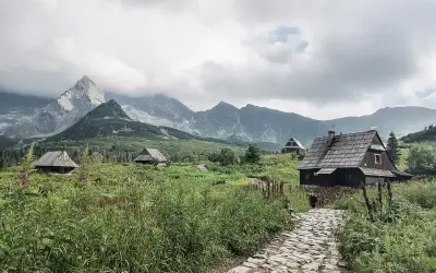 Kamienna ścieżka prowadzi do drewnianych chat pośród zieleni. W oddali widać majestatyczne góry za granicą, spowite chmurami.