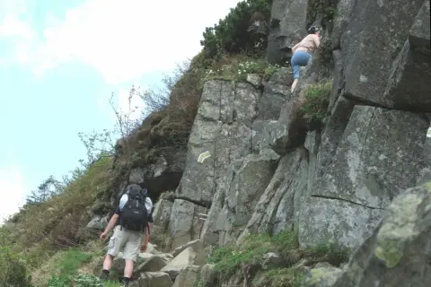Turysta z plecakiem wspina się po skalistej ścieżce, obok wspina się inna osoba.