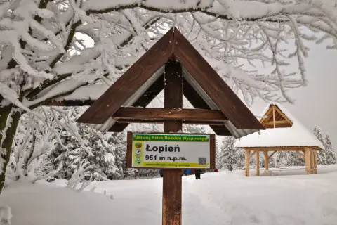 Zimowe atrakcje Beskidu Wyspowego: tablica na szczycie Łopień (951 m n.p.m.) i drewniana wiata pośród ośnieżonych drzew.