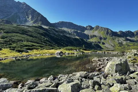 Spokojne jezioro w górach, otoczone kamieniami i zielonymi zboczami, odbija błękitne niebo.
