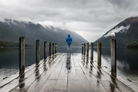 Osoba w niebieskiej bluzie stoi na moktym pomoście, patrząc na jezioro i góry. Nawet gdy pada, można podziwiać widoki.