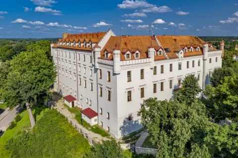 Biały zamek z czerwonym dachem, otoczony zielenią, to jeden z urokliwych zamków hoteli w Polsce.