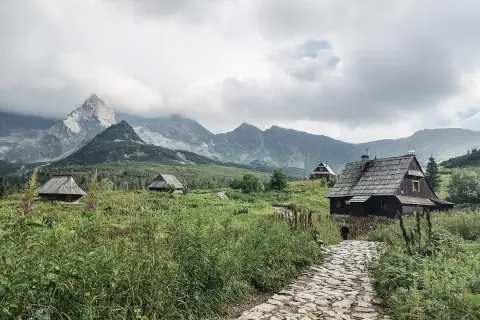 Kamienna ścieżka prowadzi do drewnianych chat pośród zieleni. W oddali widać majestatyczne góry za granicą, spowite chmurami.