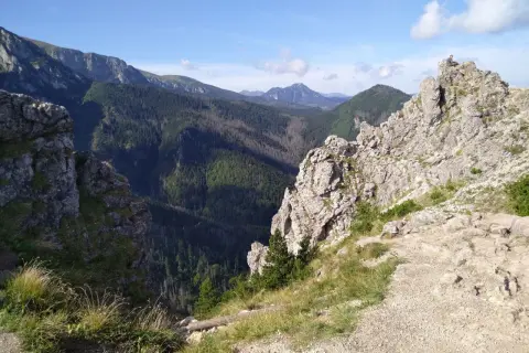 Widok na skaliste szczyty i zalesione doliny Beskidu Wyspowego. Na jednym ze szczytów siedzi człowiek, podziwiając panoramę.