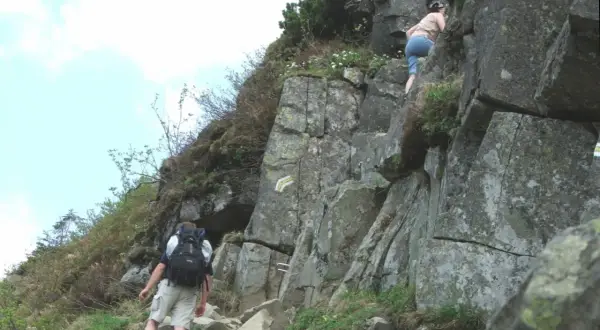 Turysta z plecakiem wspina się po skalistej ścieżce, obok wspina się inna osoba.