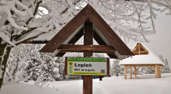 Zimowe atrakcje Beskidu Wyspowego: tablica na szczycie Łopień (951 m n.p.m.) i drewniana wiata pośród ośnieżonych drzew.