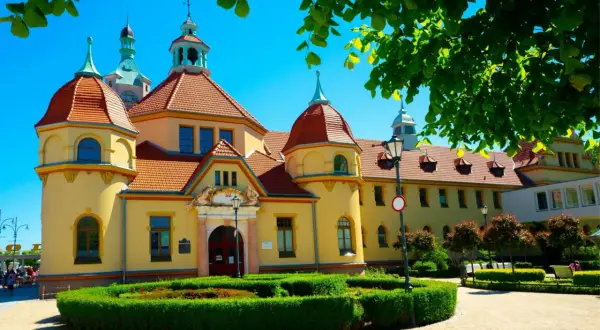 Żółty budynek z czerwonym dachem, jeden z wielu szpitale uzdrowiskowe w polsce, otoczony zielenią i drzewami.