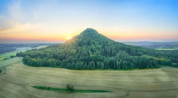 Góry Kaczawskie o wschodzie słońca. Stożkowaty, zalesiony szczyt góruje nad polami.