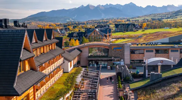 Drewniany hotel z pięknym widokiem na jesienne Tatry. W oddali widać góry i kolorowe drzewa.