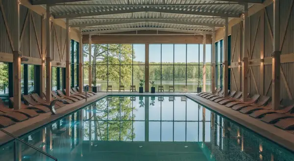 Basen w nowoczesnym spa z widokiem na jezioro. Drewniane leżaki i przeszklone ściany tworzą relaksującą atmosferę, idealną dla najlepszego spa w Polsce.