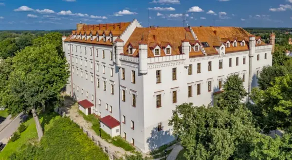 Biały zamek z czerwonym dachem, otoczony zielenią, to jeden z urokliwych zamków hoteli w Polsce.