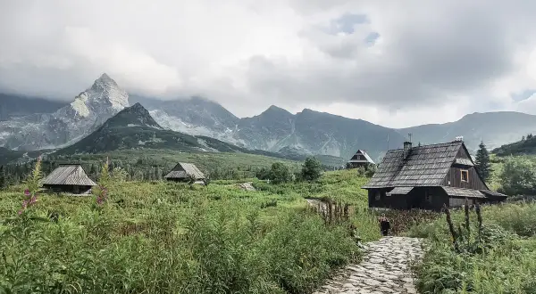Kamienna ścieżka prowadzi do drewnianych chat pośród zieleni. W oddali widać majestatyczne góry za granicą, spowite chmurami.