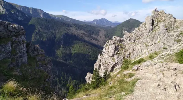 Widok na skaliste szczyty i zalesione doliny Beskidu Wyspowego. Na jednym ze szczytów siedzi człowiek, podziwiając panoramę.