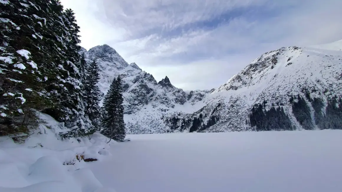 Morskie Oko zimą. Zamrożone jezioro otoczone ośnieżonymi szczytami i gęstym lasem świerkowym.