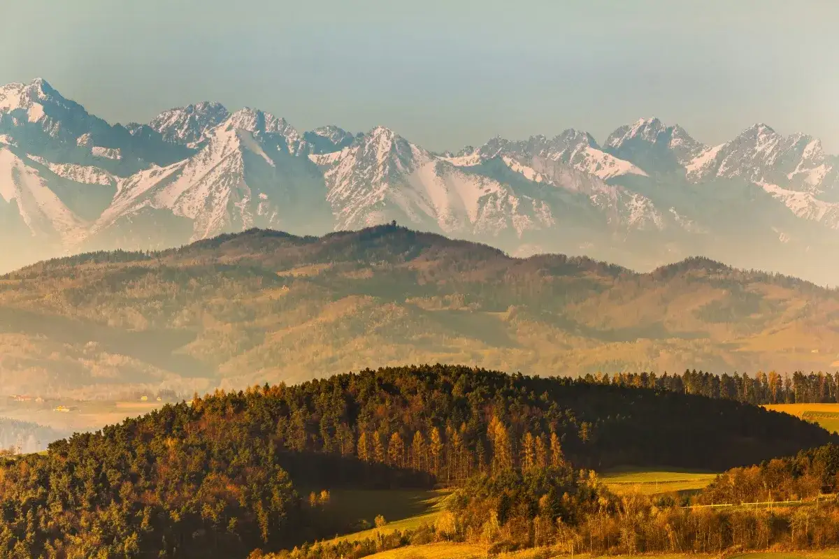 Widok na najwyższe szczyty w Polsce, Tatry, w jesiennej odsłonie. W oddali ośnieżone góry, bliżej wzgórza pokryte lasami i polami.