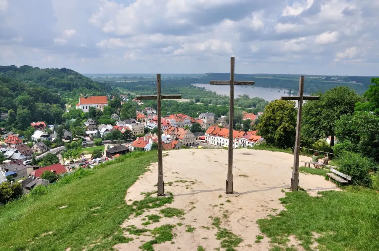 Trzy drewniane krzyże na wzgórzu górują nad miasteczkiem z czerwonymi dachami i rzeką w tle.