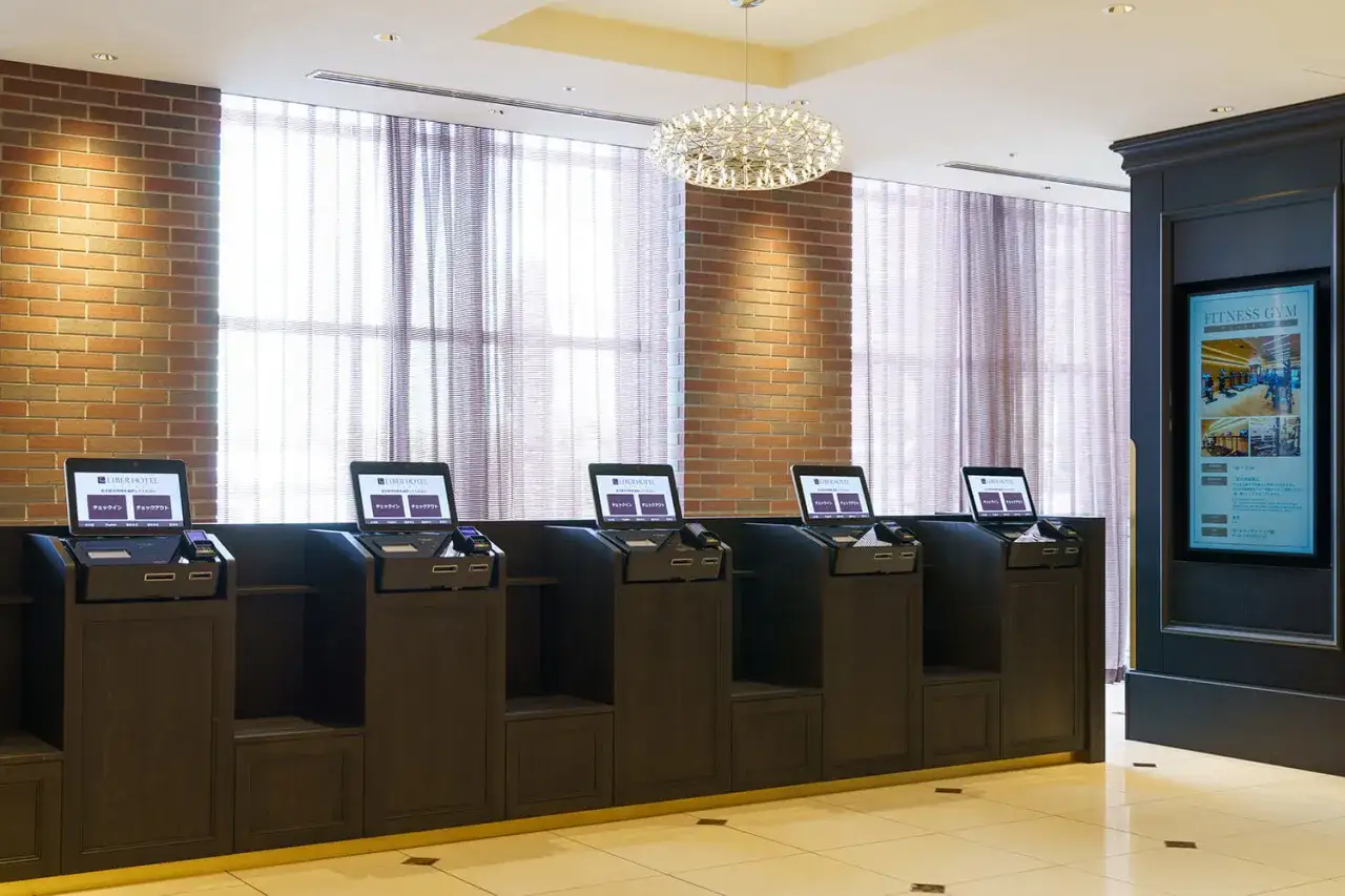 Sześć stanowisk samoobsługowych do check-in i check-out w hotelu. Eleganckie, ciemne meble i laptopy z japońskimi napisami.