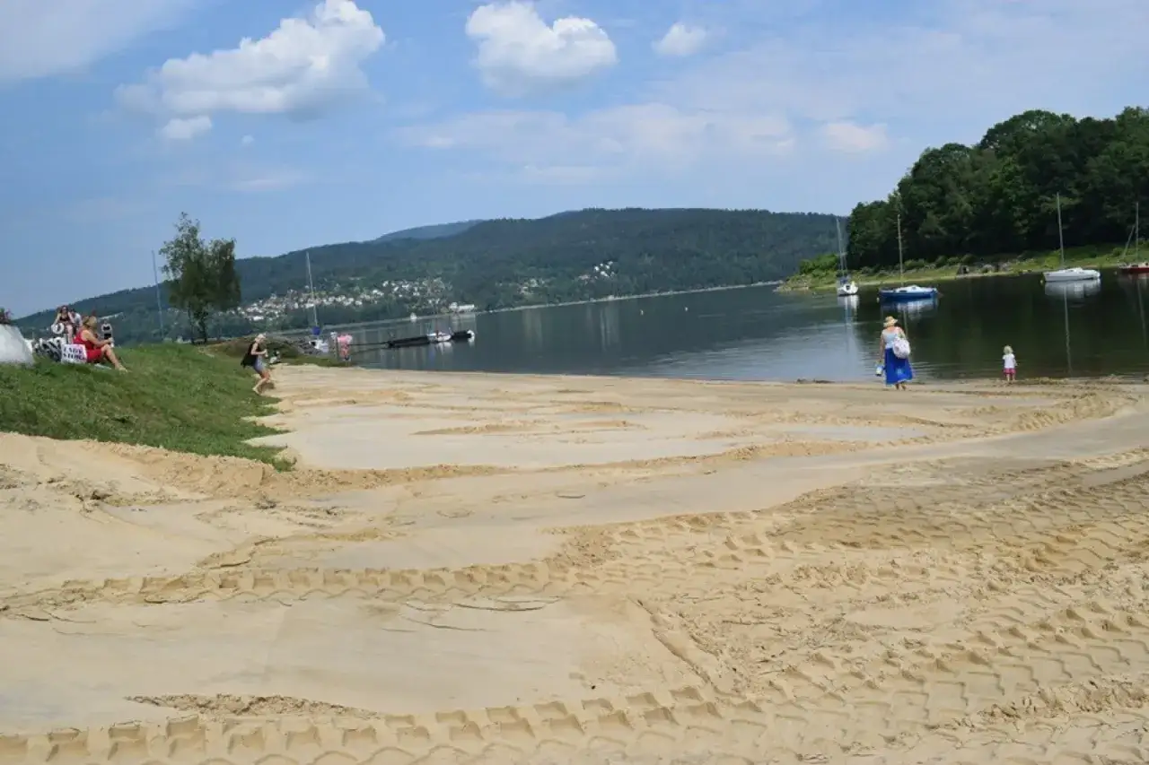 Plaża nad Jeziorem Żywieckim, z piaszczystym brzegiem i wodą, na której kołyszą się łodzie. W oddali widać zalesione wzgórza.