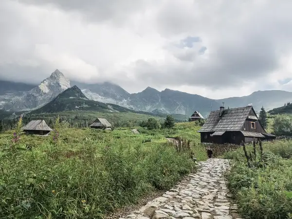 Kamienna ścieżka prowadzi do drewnianych chat pośród zieleni. W oddali widać majestatyczne góry za granicą, spowite chmurami.
