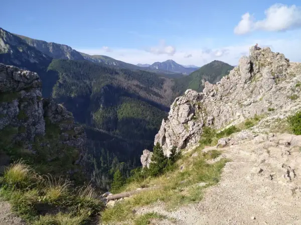 Widok na skaliste szczyty i zalesione doliny Beskidu Wyspowego. Na jednym ze szczytów siedzi człowiek, podziwiając panoramę.